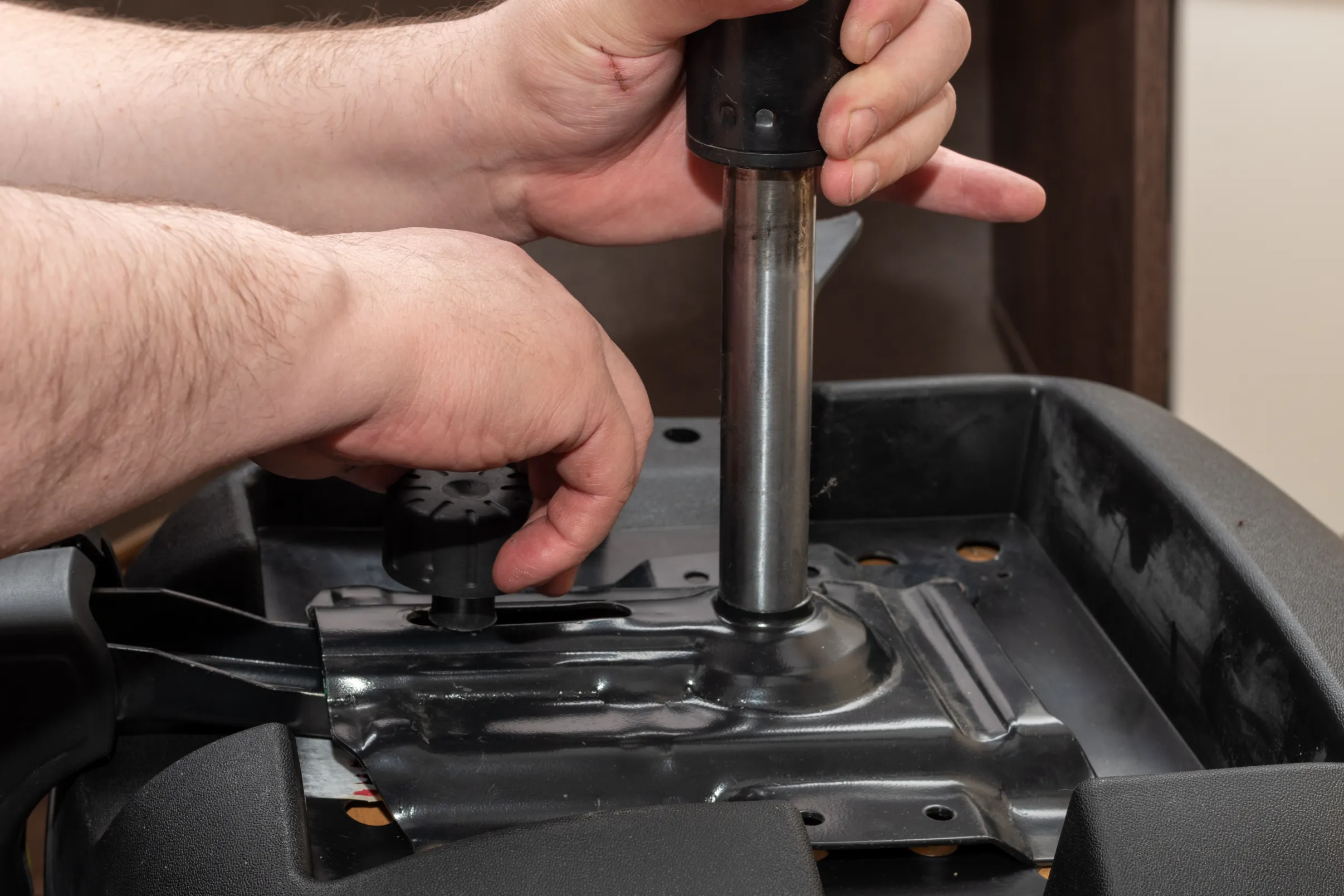 A person assembles a chair base, attaching a metal support to a black frame. Hands grip the cylinder, conveying focus and precision.
