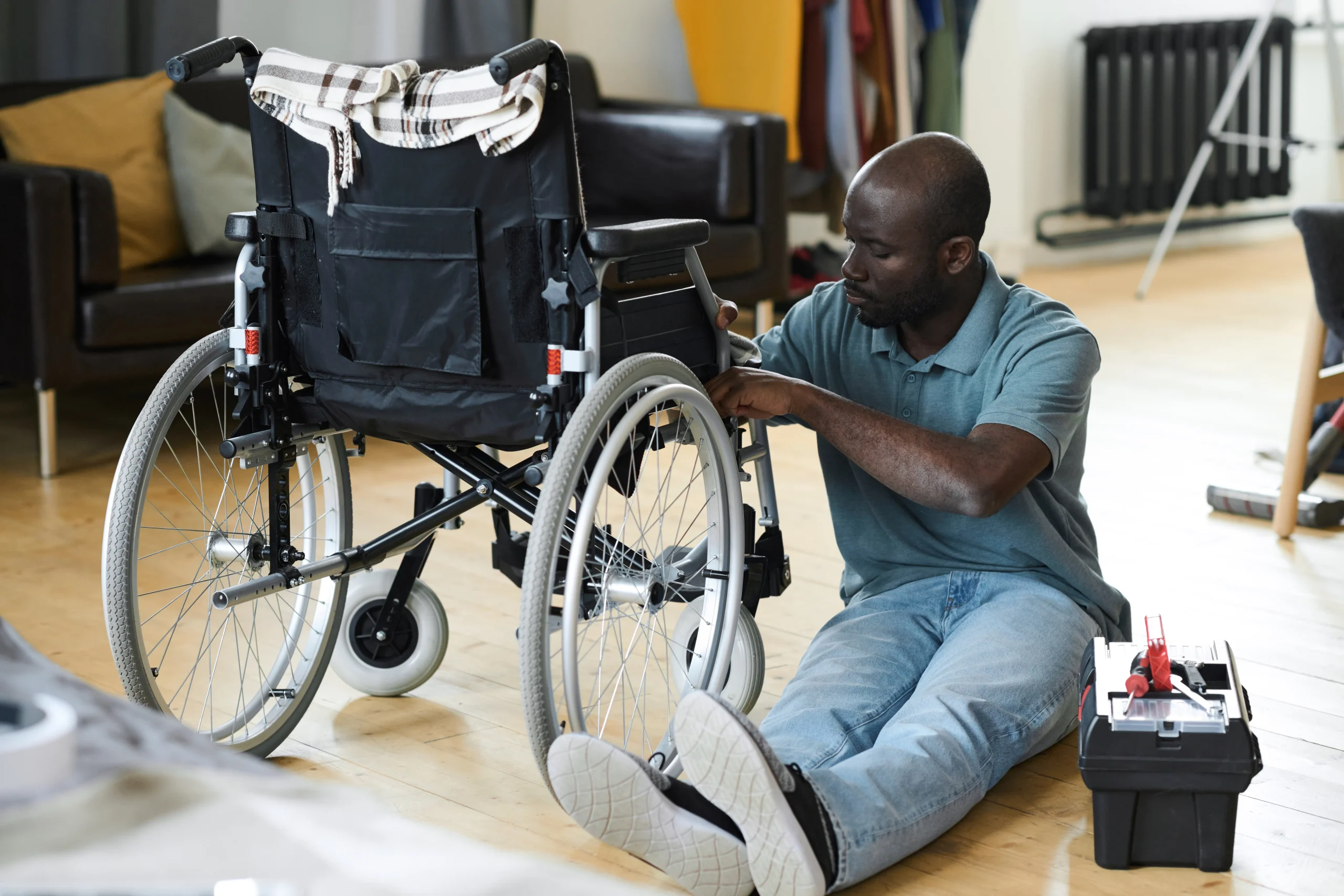 Man seated on a wooden floor repairs a wheelchair with a toolbox nearby, exuding a focused and caring atmosphere in a cozy living room.