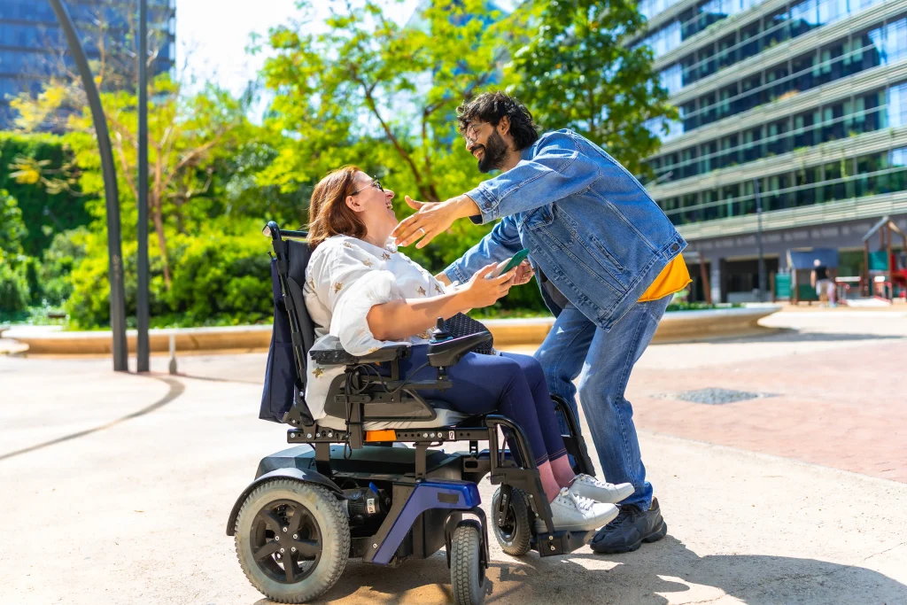 A woman in a wheelchair joyfully reaches out to a man in a denim jacket. They are outdoors near modern buildings, surrounded by greenery.