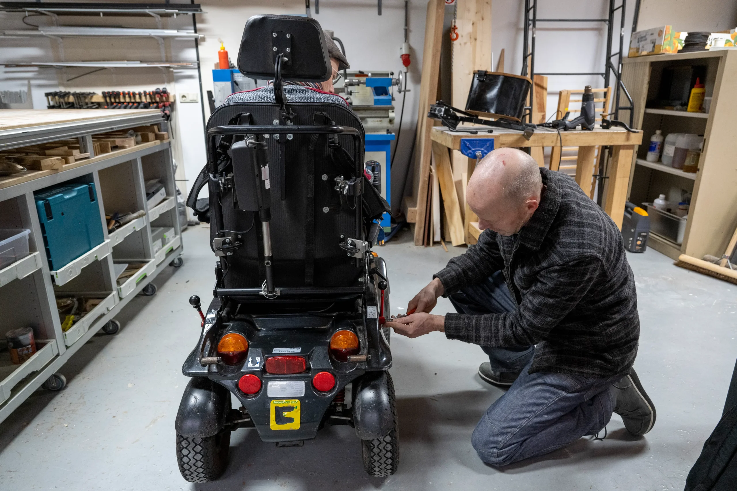 A man kneels beside a mobility scooter in a workshop, adjusting its rear light. The room is filled with tools, shelves, and wooden materials.